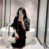 A woman in a black dress holds a white phone in front of a modern bathtub and glass partition.