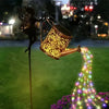 A silhouette of a fairy holding a vintage copper watering can that illuminates a trail of glowing lights on a lawn.