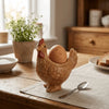A ceramic chicken egg cup with a brown, textured body sits on a wooden table next to a loaf of bread.