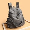 A gray soft leather backpack with metal rivets and a flap closure is displayed on a light wooden surface.