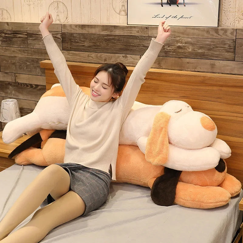 A smiling woman is lying down next to an extra-large soft dog plush pillow for snuggling and decor.