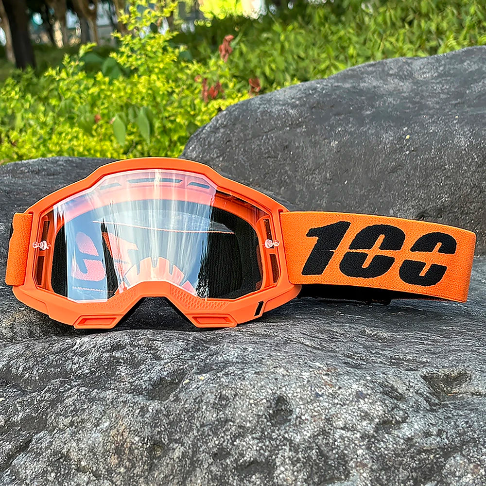 Orange motocross goggles with clear lenses resting on a dark gray rock.