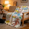 A light-colored plush throw blanket featuring colorful animal illustrations and bicycle designs draped over a wooden chair.