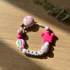 A pink silicone baby pacifier clip with a crown design and personalized name letters on a white background.