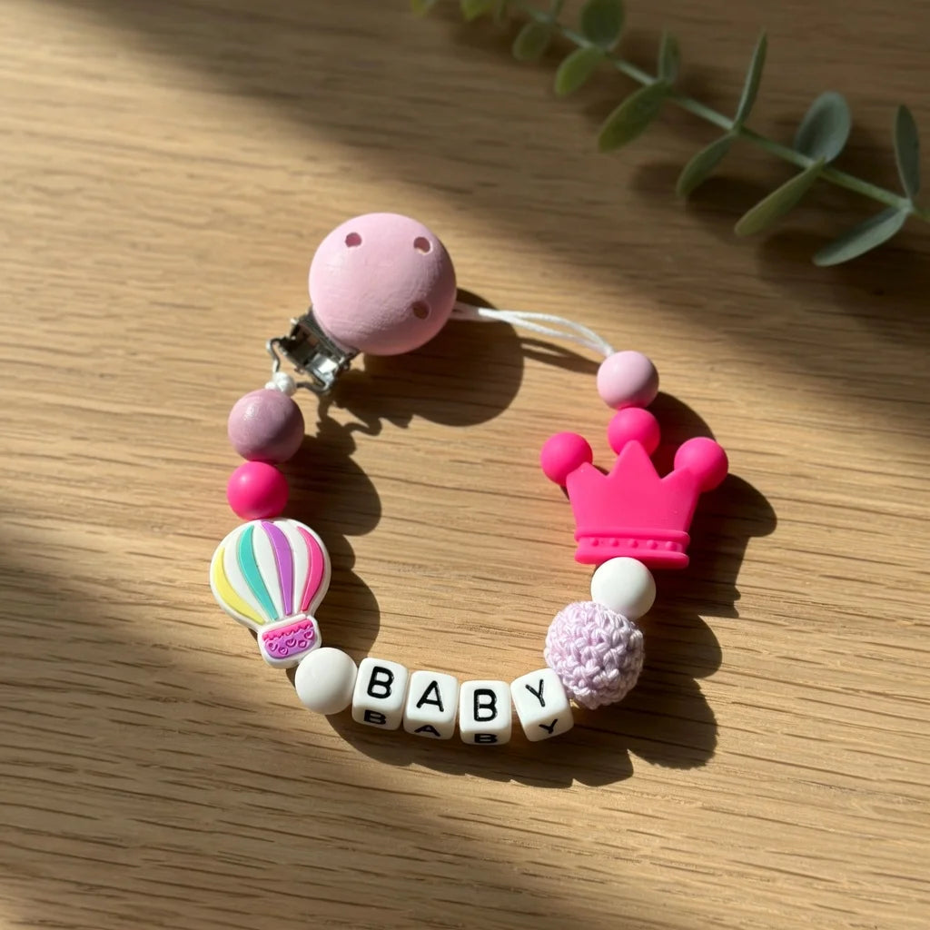 A pink silicone baby pacifier clip with a crown design and personalized name letters on a white background.