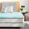 Un colchonero de espuma viscoelástica azul claro es mostrado en una cama con ropa de cama neutra y cabecero de madera.