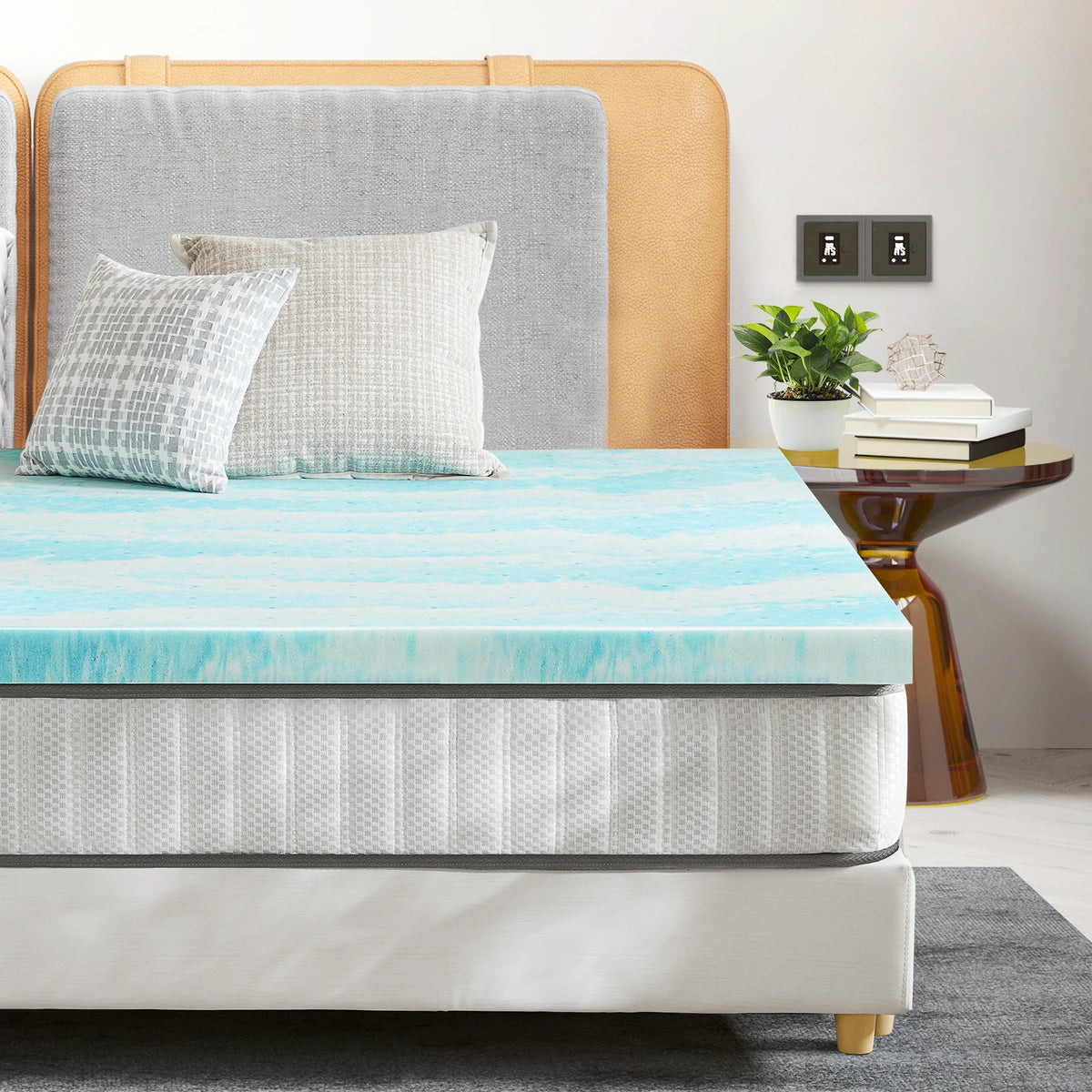 Un colchonero de espuma viscoelástica azul claro es mostrado en una cama con ropa de cama neutra y cabecero de madera.