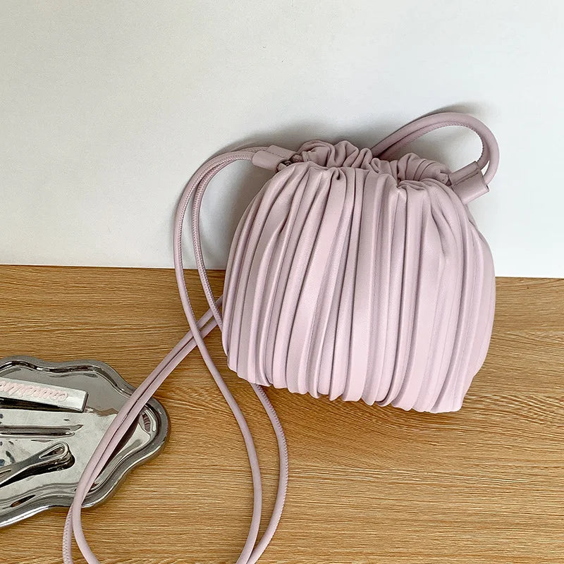 A pale pink pleated faux leather crossbody bag with a matching strap resting on a wooden surface.
