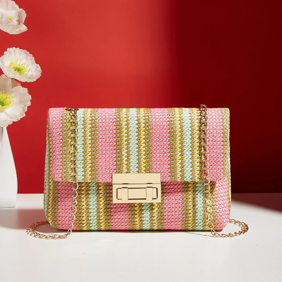 Sac bandoulière tissé rayé rose et jaune avec une chaîne dorée sur une surface blanche, fond rouge.