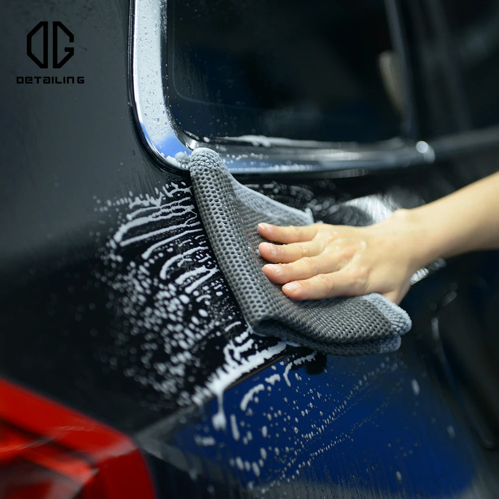 A hand is using a gray detailing towel to wipe white clay residue off a dark blue car paint surface.