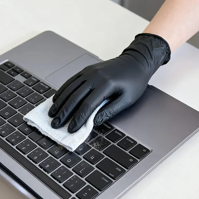 A hand wearing a black disposable nitrile glove is cleaning a laptop keyboard with a white cloth.