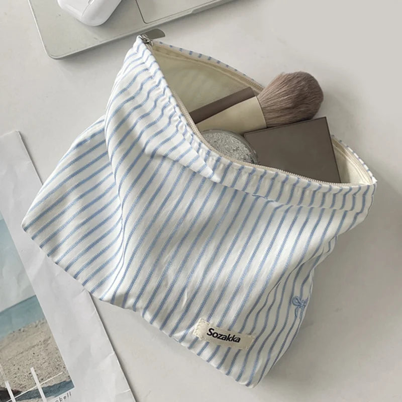 A white and blue striped makeup bag with a zipper, featuring a makeup brush and a leather product inside.