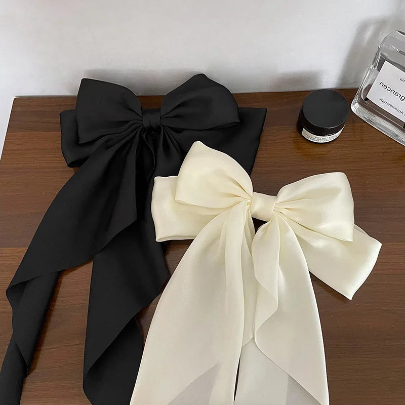 A black silk bow hair clip and a cream colored silk bow hair clip are displayed on a wooden surface.
