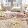 A light beige quilt with a subtle floral pattern is displayed on a bed, featuring pillows and a bedside lamp.