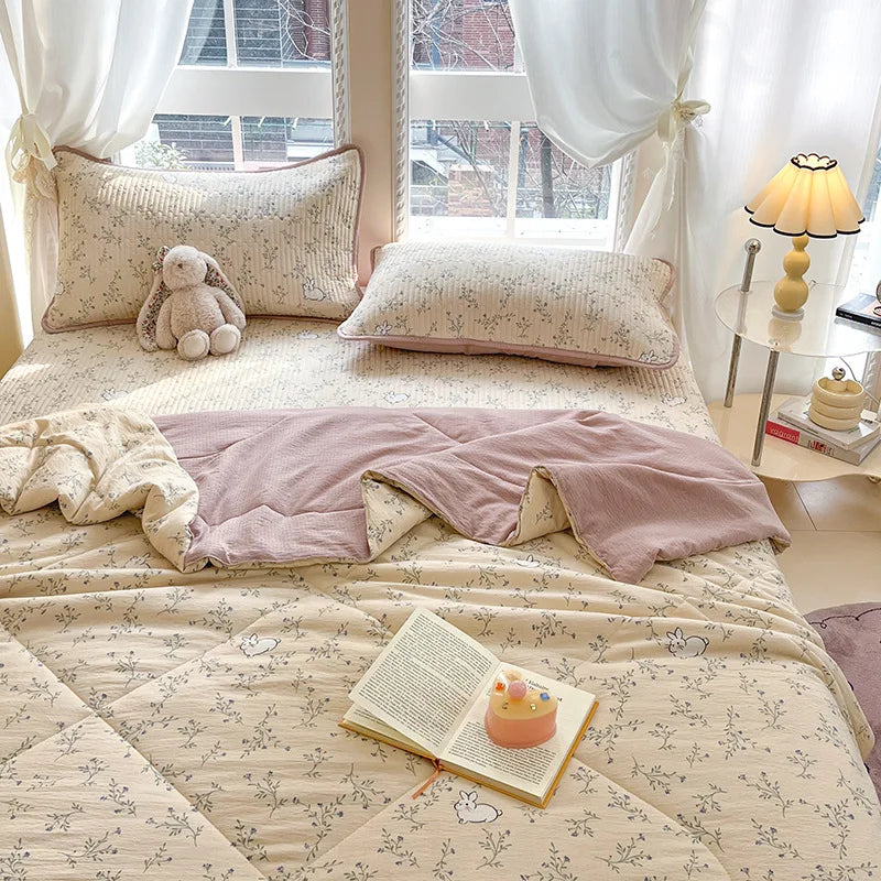 A light beige quilt with a subtle floral pattern is displayed on a bed, featuring pillows and a bedside lamp.