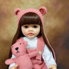 A lifelike silicone baby doll with long dark hair wearing a pink knitted outfit and a matching bear accessory.