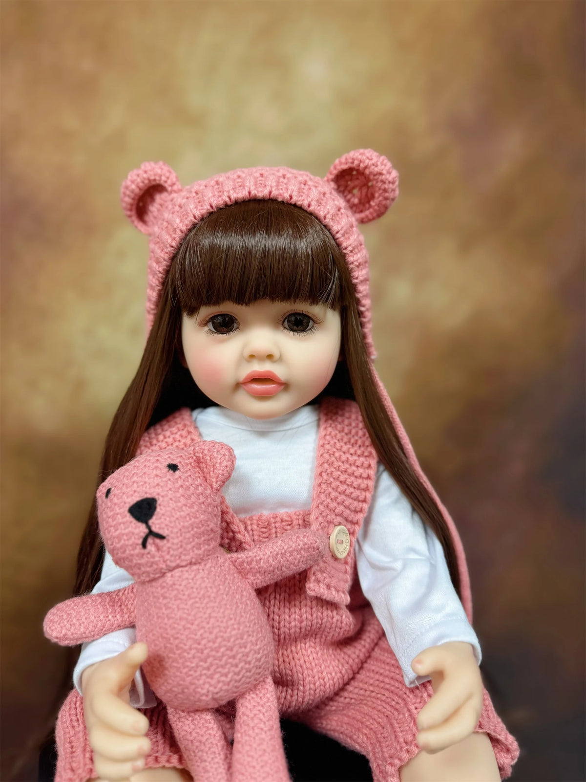 A lifelike silicone baby doll with long dark hair wearing a pink knitted outfit and a matching bear accessory.