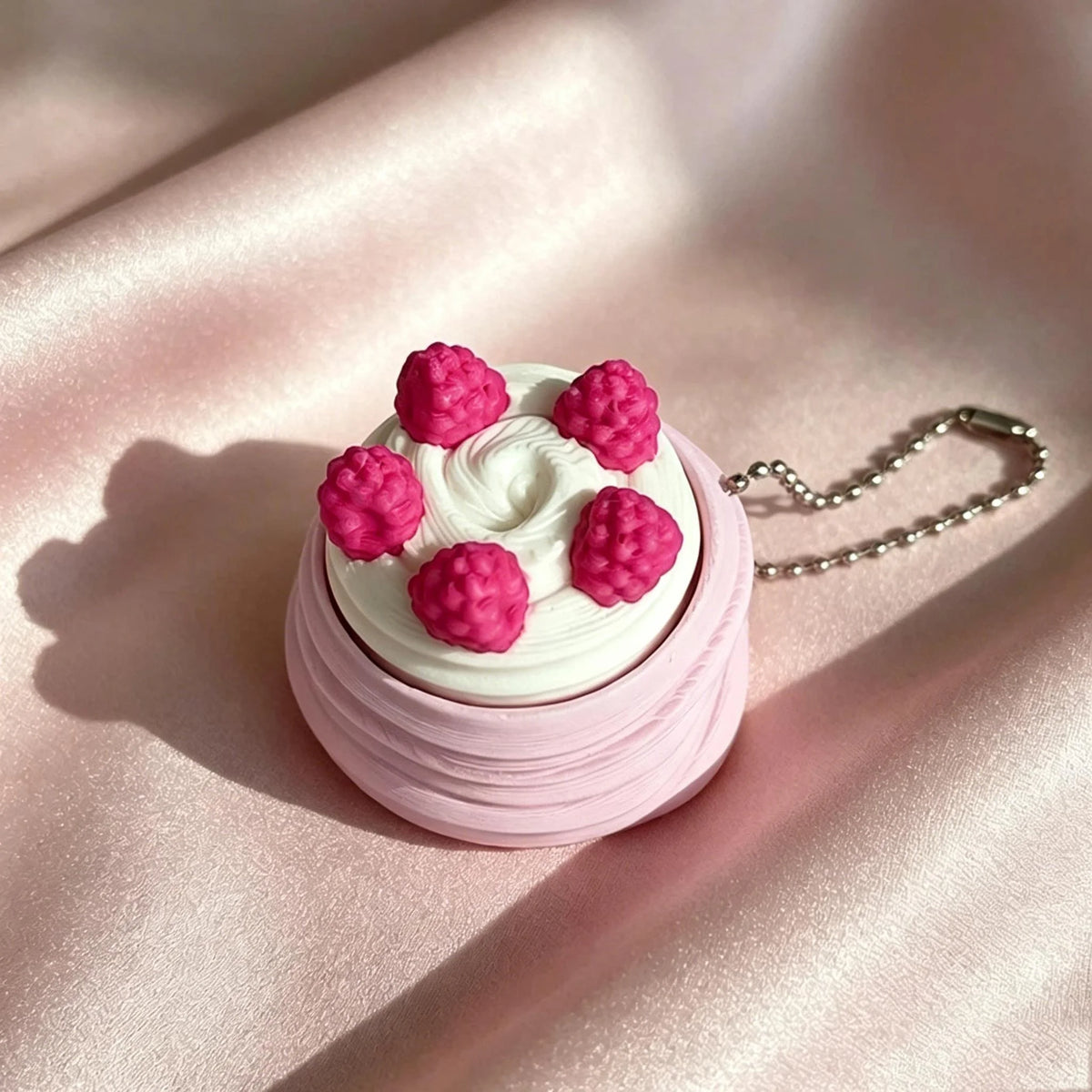 A pink frosted cake with pink meringue decorations is shown resting on a pink satin fabric with a silver chain attached.
