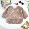 A brown waterproof long-sleeve baby feeding smock with white dinosaur pattern resting on a white table.