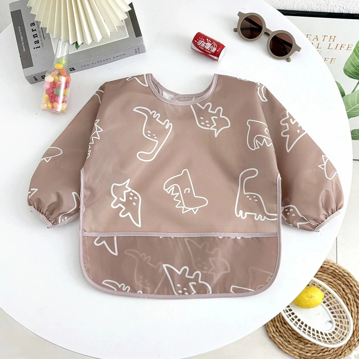 A brown waterproof long-sleeve baby feeding smock with white dinosaur pattern resting on a white table.