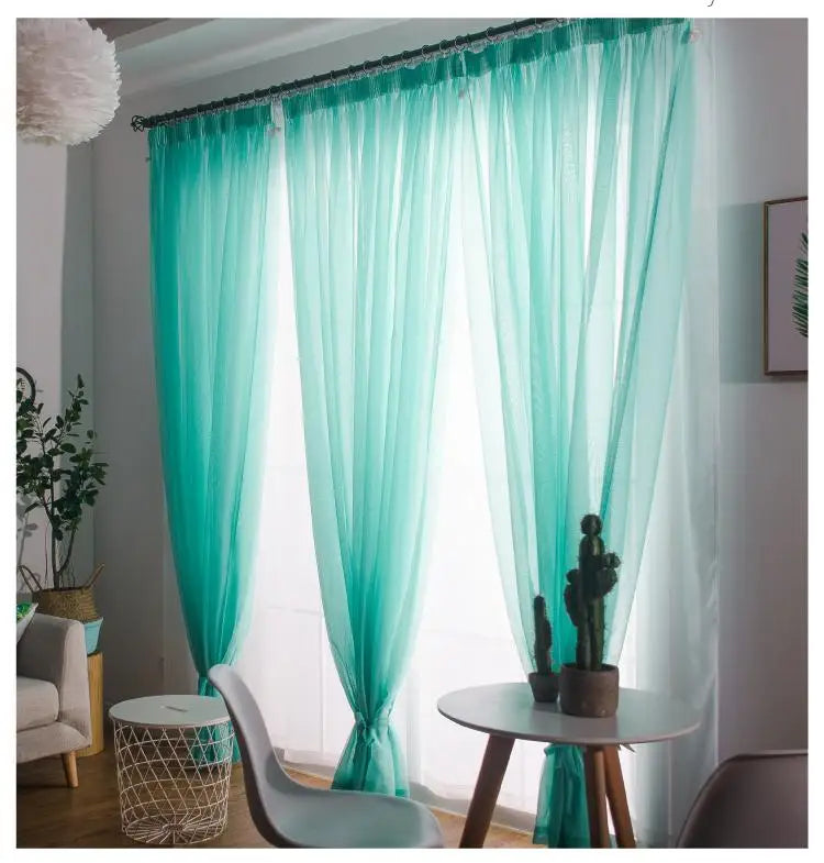 Light aqua sheer voile curtains hanging in a window with decorative greenery and a small table.