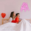 A red heart pillow rests on white bedding next to a pink neon silhouette of a hand on a wall.