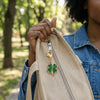 A hand holds a tan canvas bag with a gold clover leaf keychain charm against a blurred outdoor background.