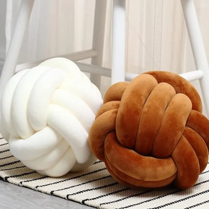 A round, hand-knotted velvet throw pillow in shades of cream and brown resting on a striped fabric.