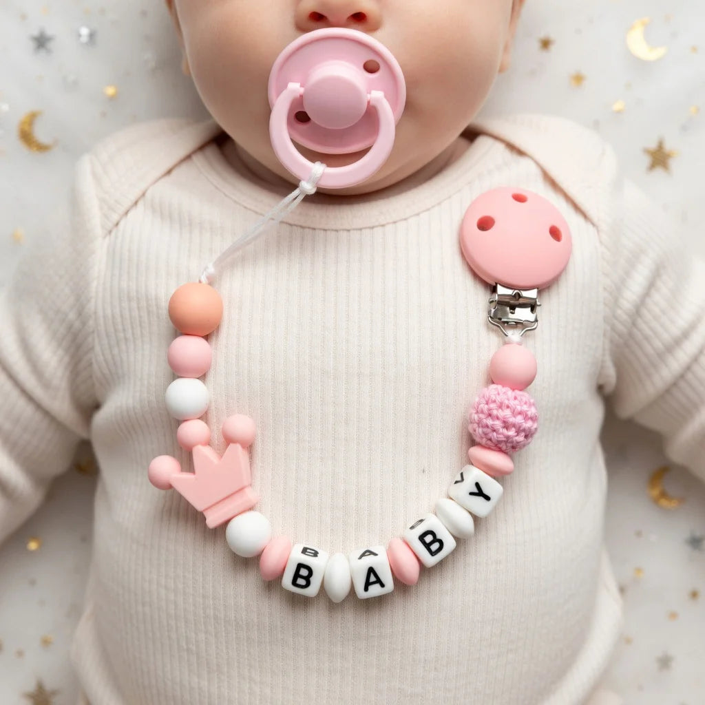 A baby wearing a pink sweater holds a personalized pacifier clip necklace with a custom name and a pink silicone pacifier.