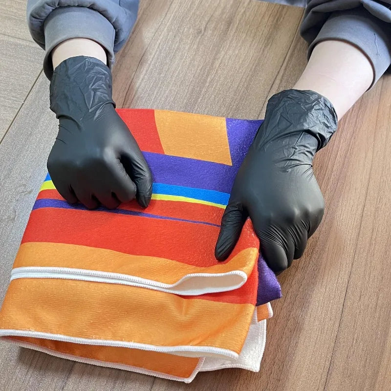 A pair of black nitrile gloves is shown wearing a stack of orange and purple disposable wipes on a wooden surface.