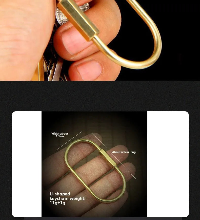 A hand holds a gold brass U-shaped keychain charm with a locking carabiner clip.