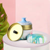 A refillable loose powder container with a blue puff and a gold lid resting on a pink surface.
