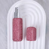 A pink glitter perfume bottle with a silver atomizer and a matching refillable glass bottle resting on a white curved surface