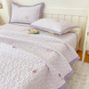 A light purple floral quilted bedspread with pink accents is displayed on a bed, featuring a matching pillow.