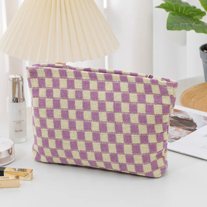 A purple and yellow plaid makeup bag with a zipper resting on a white surface next to a lamp and cosmetic items.