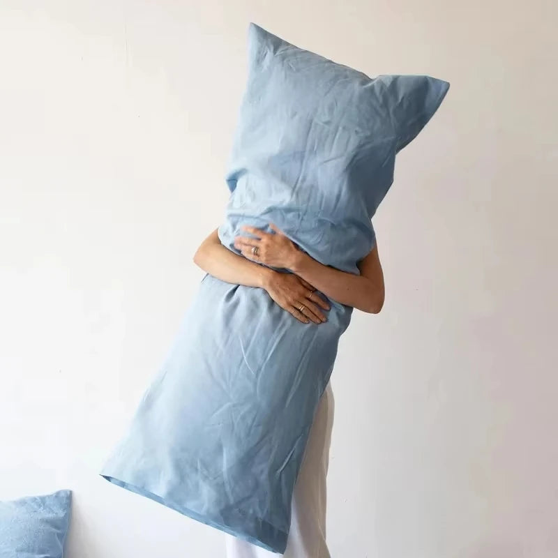 A person is holding a light blue linen pillowcase against their chest, showing the fabric and envelope closure.