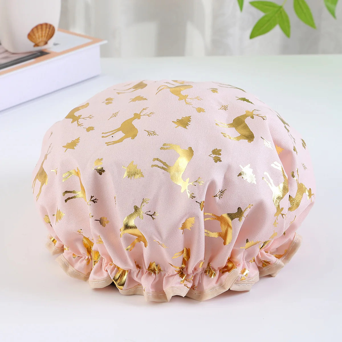 A light pink reusable elastic shower cap decorated with gold reindeer patterns.
