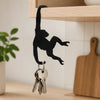 A black monkey silhouette hangs from a wooden wall hook, with keys attached below, against a light wooden shelf.