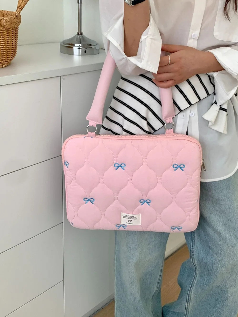 A pink quilted laptop shoulder bag with a light pink strap is being held by a person wearing a white shirt and jeans.