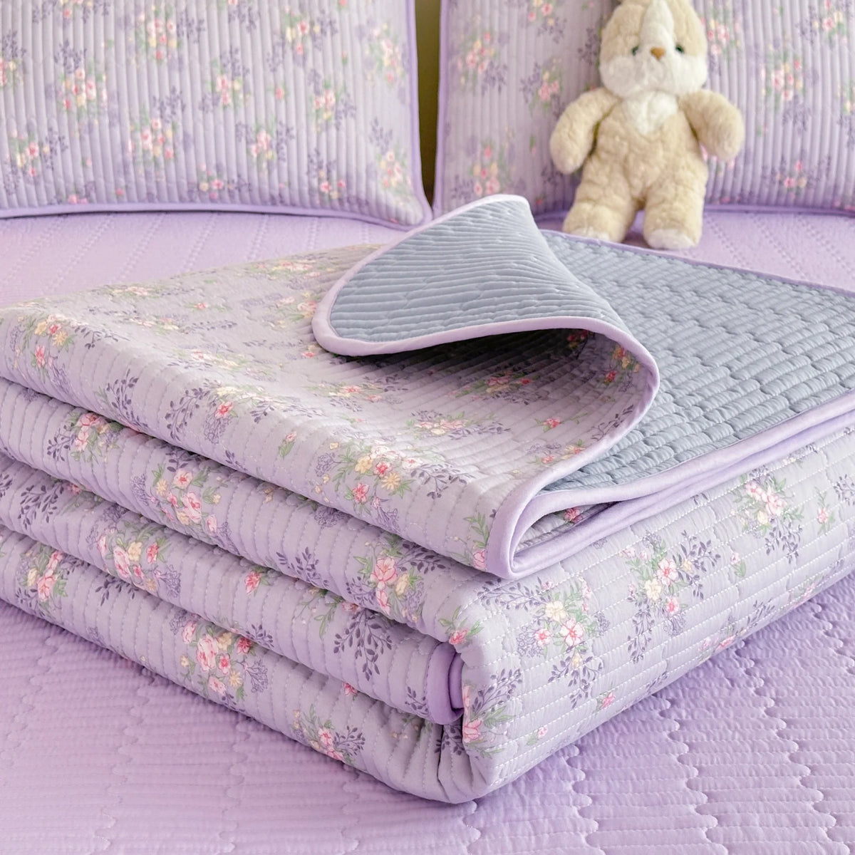 A stack of light purple floral quilted bedspreads with dust-proof covers resting on a lavender surface.