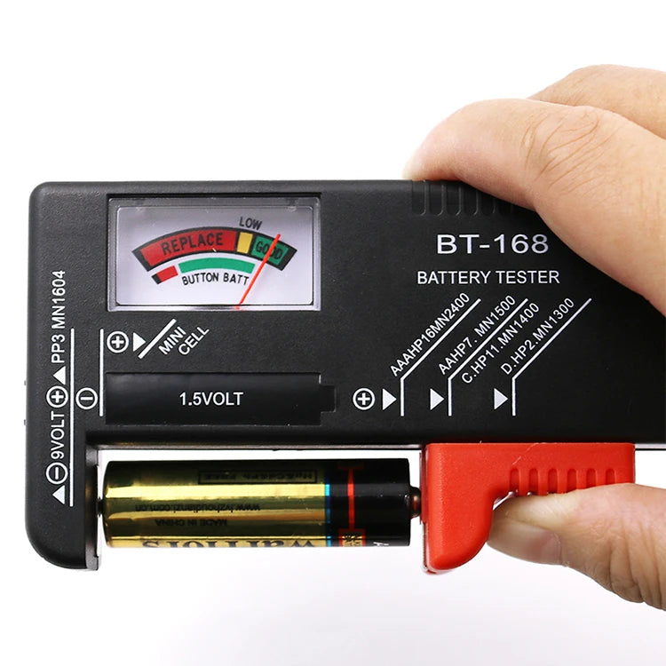 A hand holds a black battery tester device with a red indicator and a battery cell inserted.