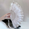 A white folding fan with lace trim and a golden edge is held by a hand against a light background.