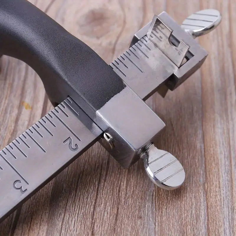 A close-up of a silver adjustable leather strap cutter with a black handle and marked blades resting on a wooden surface.