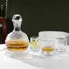 A handcrafted crystal wine decanter with a rotating aerator sits on a white table next to various drinking glasses.