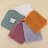 A stack of five colored cotton muslin baby burp cloths including white, green, orange, and purple.