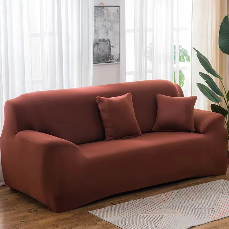 A modern, rust-colored elastic stretch sofa cover with two throw pillows placed in a bright room.