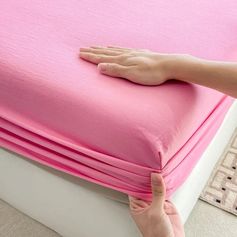 A hand is folding a bright pink fitted sheet with an elastic band on a white bed frame.