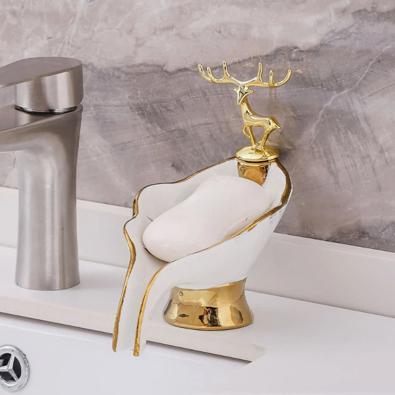A gold deer figurine sits atop a white leaf-shaped ceramic soap dish with a drain rack.