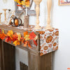 Linen table runner featuring an autumn pattern of pumpkins and maple leaves displayed on a wooden mantelpiece.