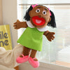 A hand puppet of a Black woman with curly hair and bright green dress standing in front of a window.